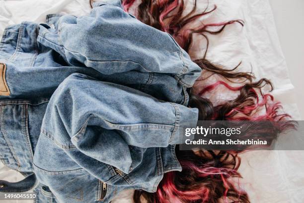 young woman with beautiful wavy highlighted hair, thick black bang and pink strands lies on white texture fabric. attractive brunette girl in denim jacket hides her face with her hands. - thick white women ストックフォトと画像