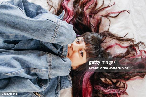 young woman with beautiful wavy highlighted hair, thick black bang and pink strands lies on white texture fabric. attractive smiling brunette girl peeking out from under denim jacket. - thick white women ストックフォトと画像