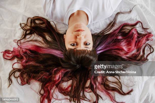 attractive brunette girl in white t-shirt lies on white textured fabric. close-up of female portrait of young woman with beautiful long curly highlighted hair, thick black bang and pink strands. - hair colour stock pictures, royalty-free photos & images