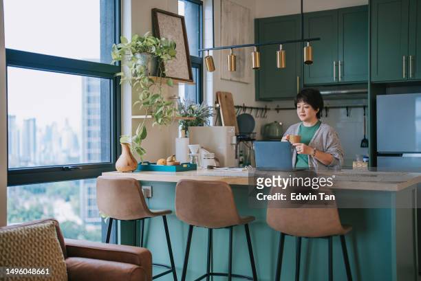 asian mature chinese woman working at home at kitchen counter using laptop - color enhanced stock pictures, royalty-free photos & images