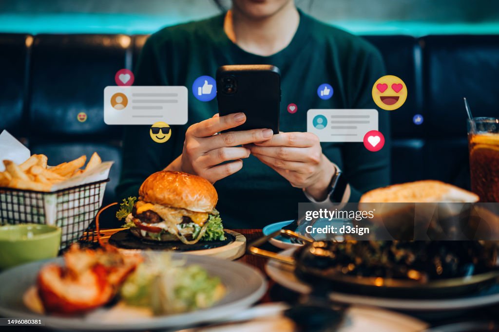 Cropped shot of young Asian woman using smartphone on social media app during meal in restaurant. Viewing posts, giving like, love, comment, making friends on pages in the city. People engaging in networking with technology. Social media addiction concept