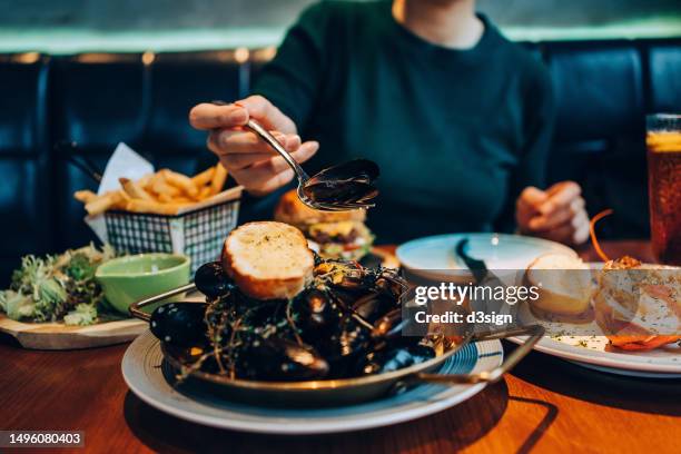 mid-section of young asian woman enjoying lunch in a seafood restaurant. she is eating mussels with white wine sauce, with assorted dishes freshly served on the dining table. people, food and lifestyle - foodstyling stock-fotos und bilder