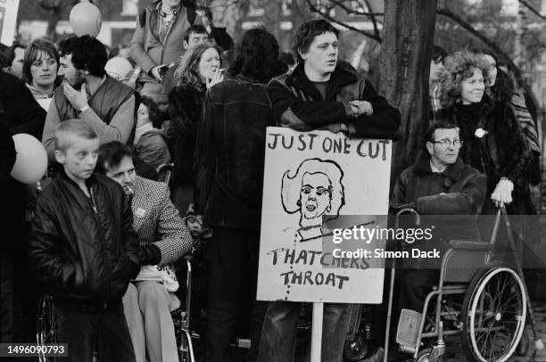 Man among a crowd of protesters leans on a placard titled 'Just One Cut - Thatcher's Throat' during an anti-cuts march, February 4th 1981.