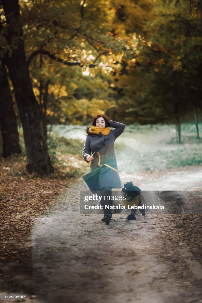 Embracing spirit of fall: radiant plus-size woman in coat and hat shares laughter and fun with her black poodle dog, creating lasting memories in serene backdrop of an autumn park