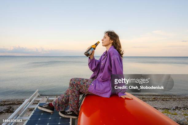 woman sitting on roof of camper van and drinking beer near the sea - zonne eiland stockfoto's en -beelden