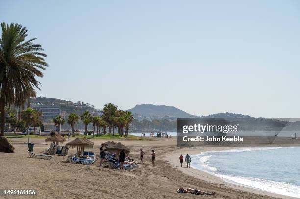 People walk along and sunbathe on Playa La Caleta beach by sunbeds on a sunny clear blue sky day on April 20, 2023 in Malaga, Spain.