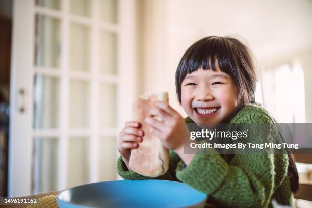 lovely cheerful girl smiling joyfully at the camera while enjoying sandwich at home in the morning - child food stock pictures, royalty-free photos & images