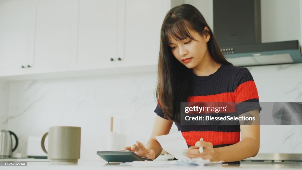Young Asian woman sorting out payment bills and household finances in the kitchen, inflation and financial crisis concept