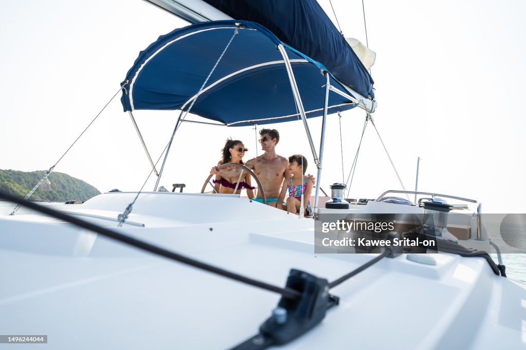 Caucasian happy family driving yacht outdoors on the sea during summer. Young beautiful couple in bikini hanging out and spending time with daughter sailing boat during holiday vacation trip together.