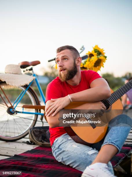 young hipster guy holding acoustic guitar while sitting on dock in nature. summer and travel concept. - classical guitarist stock pictures, royalty-free photos & images