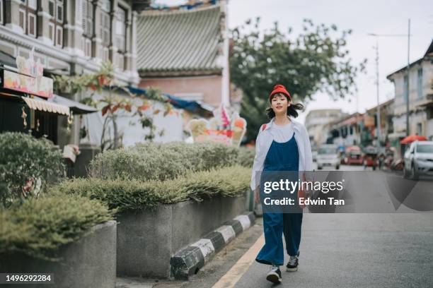 asiatische chinesische junge frau mit lätzchen, die in der altstadtstraße von penang mit skateboard spazieren geht - jeans latzhose frau stock-fotos und bilder