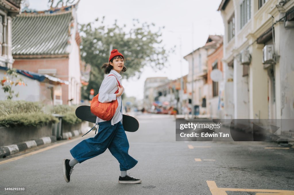 Glückliche asiatische chinesische junge Frau, die Straße mit Skateboard in der Altstadt überquert