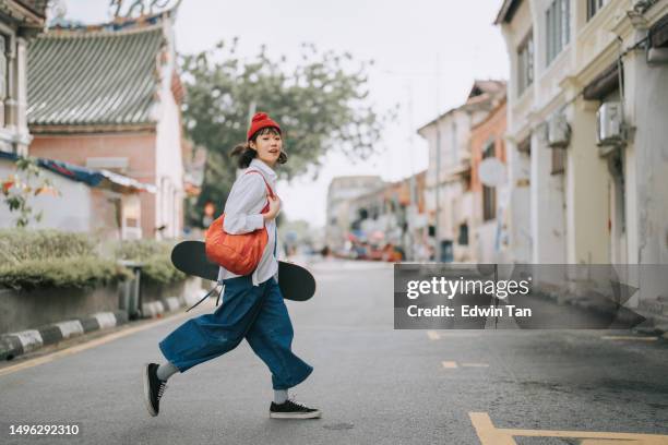 happy asian chinese young woman crossing road carrying skateboard in old town - urban fashion stockfoto's en -beelden