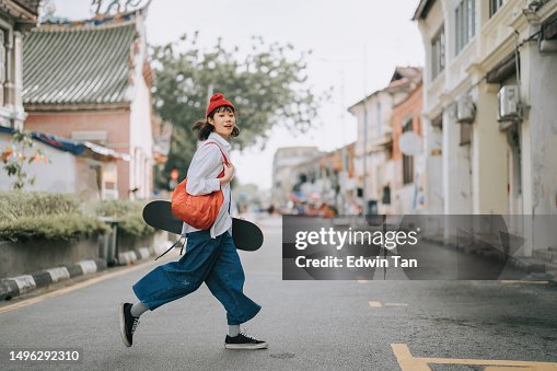 Happy Asian Chinese young woman crossing road carrying skateboard in old town Happy Asian Chinese young woman crossing road carrying skateboard in old town
