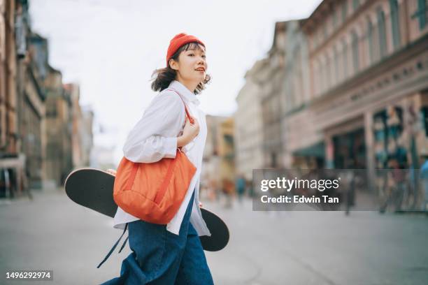 feliz joven china asiática cruzando la carretera llevando monopatín en el casco antiguo - monopatín actividades recreativas fotografías e imágenes de stock
