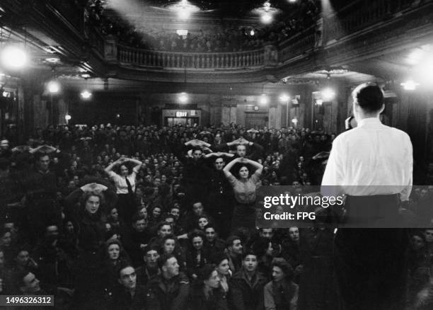 British hypnotist Peter Casson, on stage with back to the camera, conducts a mass hypnosis event, with members of the audience standing with their...