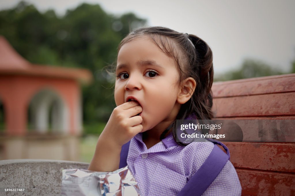 Cute schoolgirl sitting on park bench and she eating snack after school