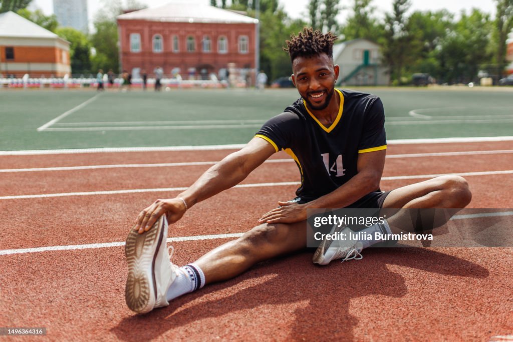 Cheerful Gen Z professional sportsman of Iranian ethnicity stretching leg muscles while siting on ground at sports stadium