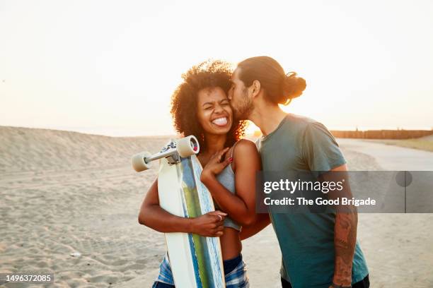 man kissing girlfriend holding skateboard while standing at beach - couple tongue kissing stock-fotos und bilder