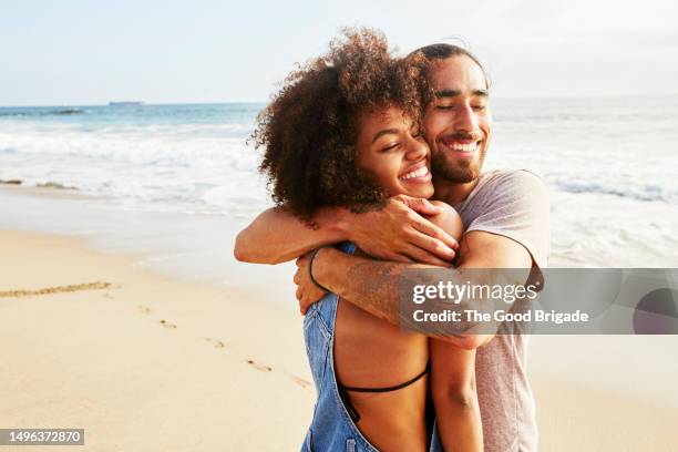 smiling boyfriend with eyes closed embracing girlfriend at beach - fonds marins foto e immagini stock