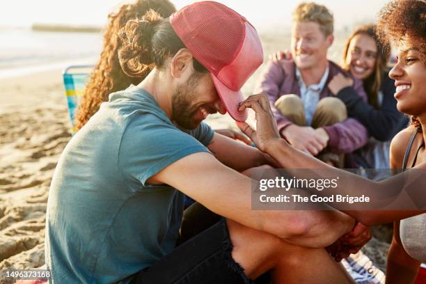 smiling woman adjusting cap of boyfriend sitting at beach - trucker hat stock pictures, royalty-free photos & images