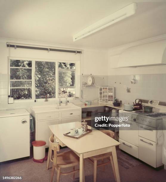 View of an early Sixties bungalow kitchen interior with gas cooker, sink, tiled walls, corner unit for pan storage, toploader washing machine, table...