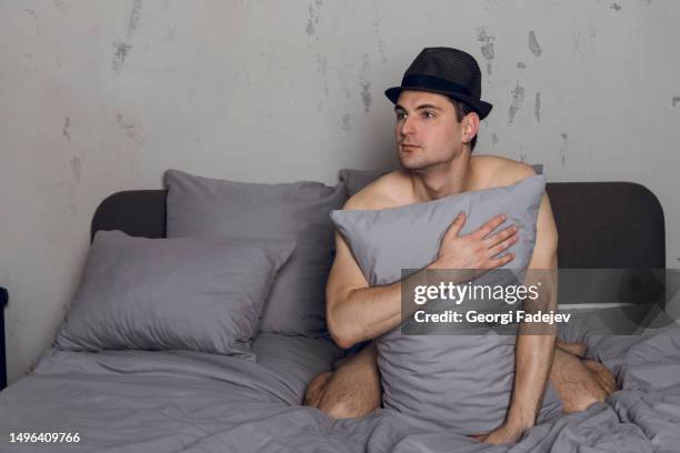 handsome slim young man in hat covers himself with a pillow on the bed. a guy covering his body with the pillow. - caught cheating stockfoto's en -beelden