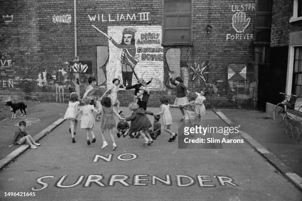 Children playing 'Ring a Ring o' Roses' on a street marked with graffiti reading 'No Surrender', Belfast, Northern Ireland, 1962.