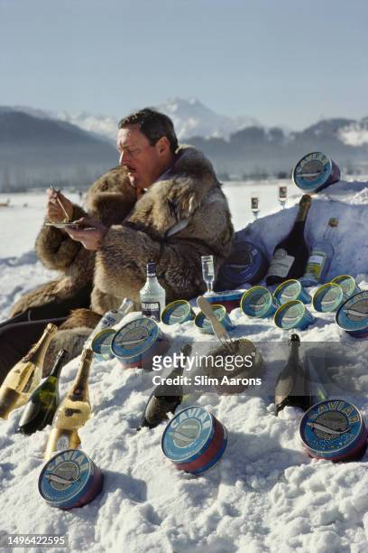 Man wearing a fur coat sits in the snow surrounded by caviar, vodka, and champagne, St. Moritz, Switzerland, 1989.