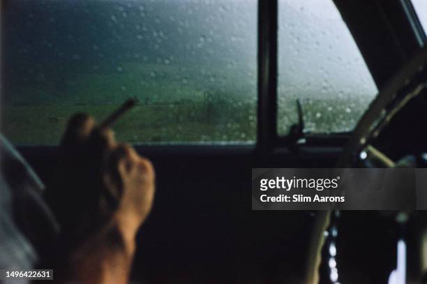Close-up defocussed shot showing the hand of the photographer Slim Aarons holding a cigarette in a car with raindrops on the window, 1959.