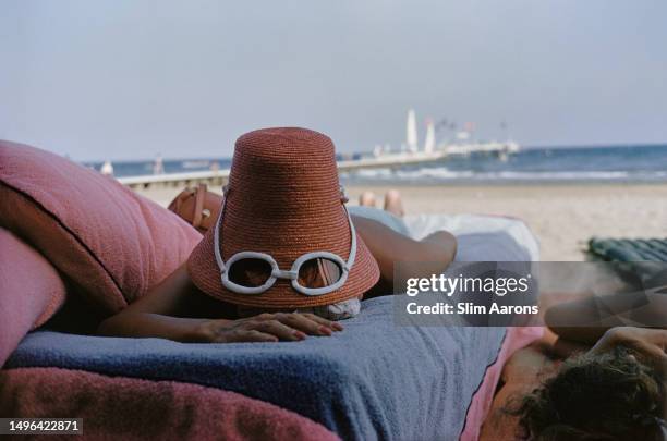 Woman sunbathes wearing a novelty straw sun hat with built-in sunglasses, Venice, 1954.