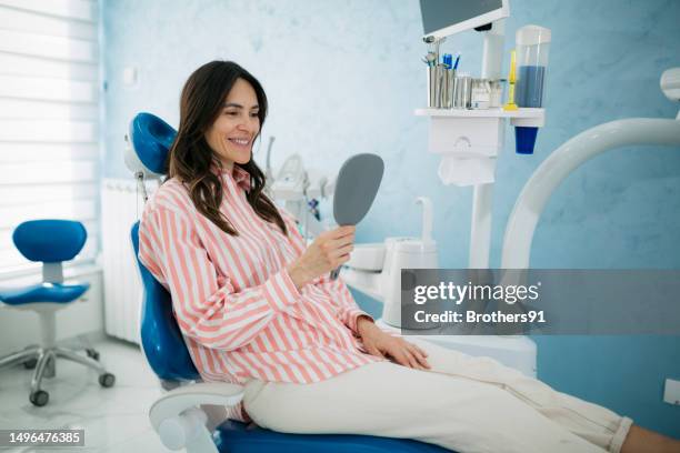 mature woman patient looking into hand held mirror and smiling at dental clinic - implante dentário imagens e fotografias de stock