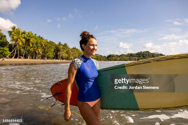 a woman with a colorful surfboard walking towards the ocean. - san jose costa rica stock pictures, royalty-free photos & images
