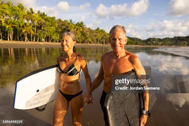 a senior couple holding boogy boards ready for a fun day in the ocean. - active seniors surfing stock pictures, royalty-free photos & images