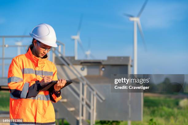 asian female wind turbine engineer wearing a hard hat checking the quality of wind turbine with a digital tablet against a wind turbine in the wind mill yard. - climate scientist stock pictures, royalty-free photos & images