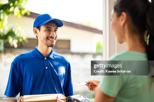 asian young delivery man delivering package to female customer at home. attractive postman in blue uniform working, deliver a box to woman then receive credit card payment infront of the door in house - express delivery stock pictures, royalty-free photos & images