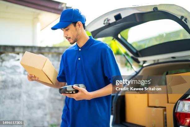 asian male delivery in blue uniform handling a box of parcel from car. attractive young postman worker express and carry a ordering box of products then giving to a customer in front of the house. - material handling stock pictures, royalty-free photos & images