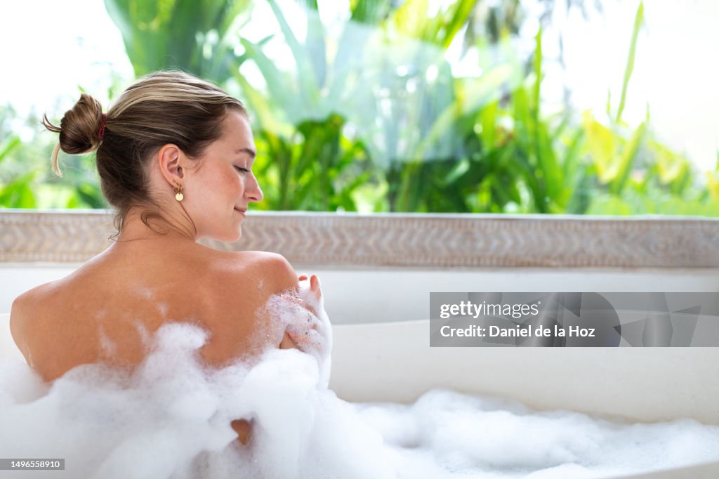 Rear view of young caucasian woman enjoying bubble bath. Female embracing herself while taking relaxing bath in tropical paradise hotel. Copy space.Wellness