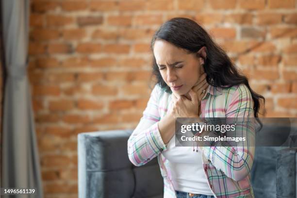 young woman suffering from thyroid disorder, holding her throat, sitting in pain at home on sofa. - menselijke mond stockfoto's en -beelden