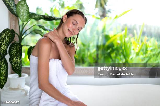 young caucasian woman wrapped in white cotton towel giving herself neck and shoulder massage after relaxing bubble bath in tropical resort hotel. copy space. self care - bali massage ストックフォトと画像