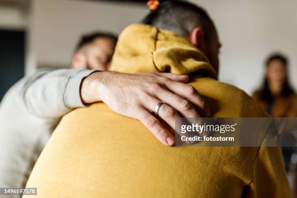 uomo che abbraccia il suo amico che sta condividendo la sua storia alla sessione di terapia di gruppo - group of patients foto e immagini stock