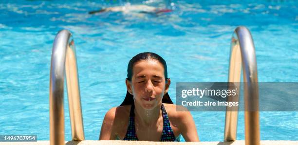 close-up of front view of a cute smiling girl in a swimsuit climbs the ladder to get out of the pool after enjoying a swim against the blurred background of the swimming pool - portrait blurred background stockfoto's en -beelden