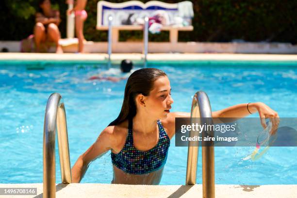 close-up of front view of a cute smiling girl in a swimsuit climbs the ladder to get out of the pool after enjoying a swim against the blurred background of the swimming pool - portrait blurred background stockfoto's en -beelden