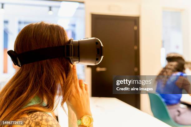 seated woman with long hair and a tattoo on her shoulder with virtual reality glasses and another woman in front seated with virtual reality glasses - looking over his shoulder stockfoto's en -beelden