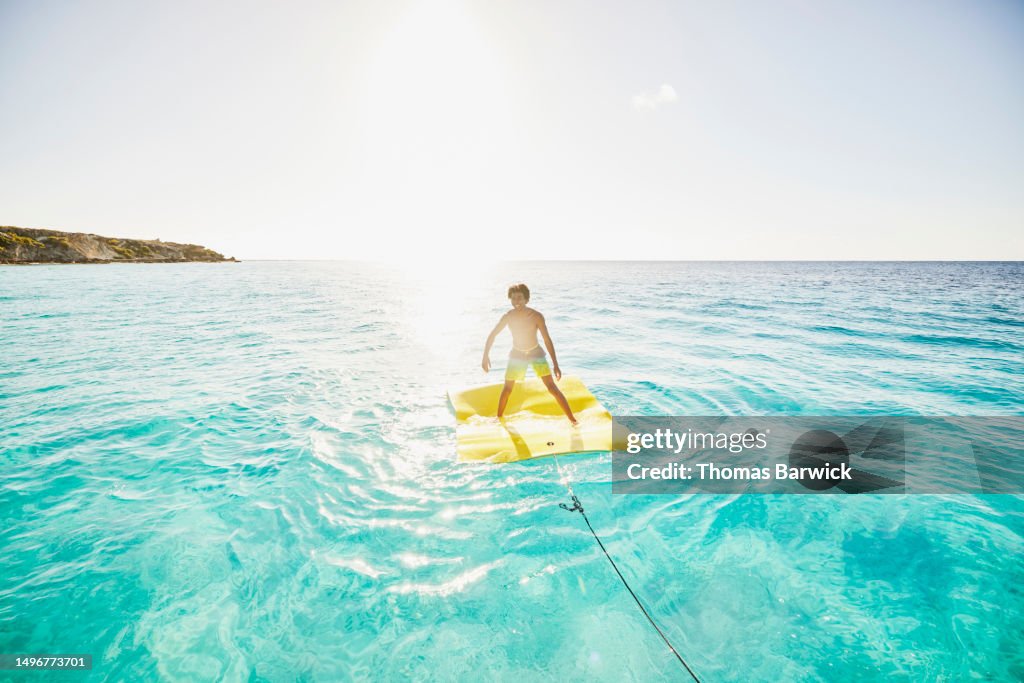 Extreme wide shot smiling teenage boy balancing on swim float in ocean