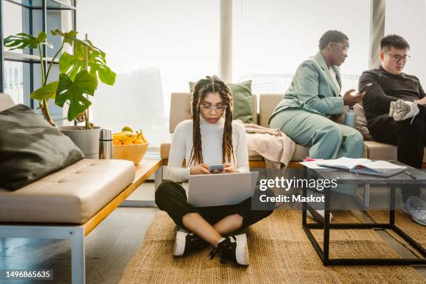 programmer using mobile phone while sitting with laptop on carpet in creative office - carpet icon stock pictures, royalty-free photos & images