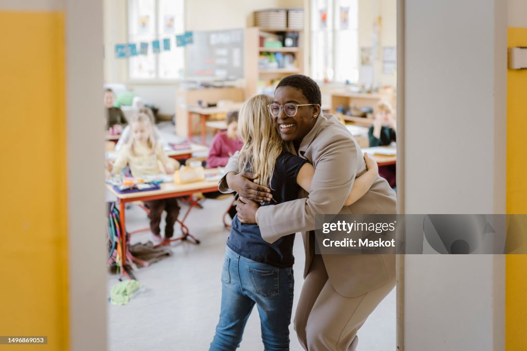 Smiling female teacher and schoolgirl embracing each other at doorway in classroom