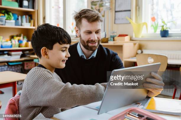 smiling male teacher assisting schoolboy using tablet pc at desk in school - male teacher in a classroom stock-fotos und bilder