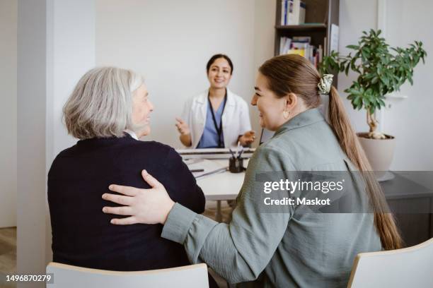 rear view of smiling daughter with hand on back of mother sitting in clinic - indian old woman stock-fotos und bilder