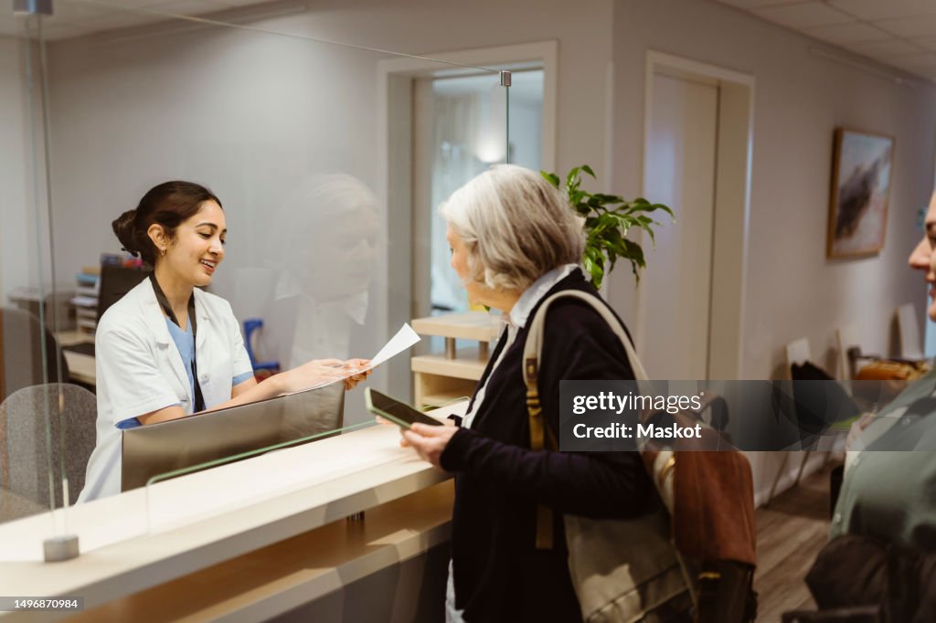 Smiling receptionist explaining form filling procedure to patient through transparent shield in clinic